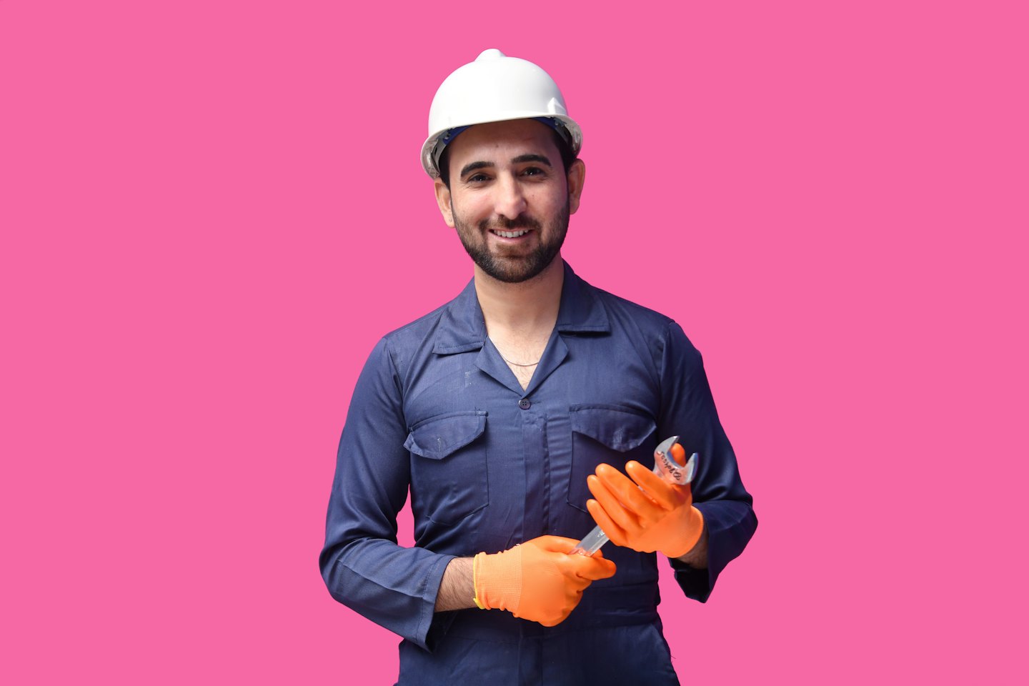 A man in a blue coverall and white hard hat holds a wrench.
